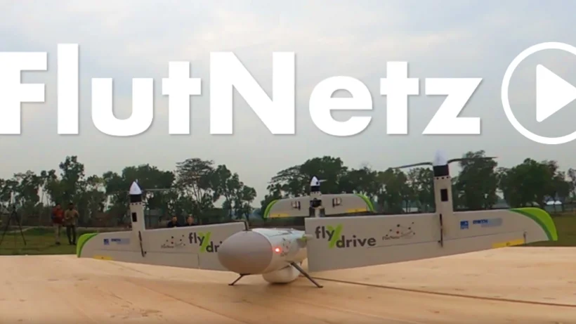 Eine unbemannte FlutNetz-Drohne mit der Aufschrift „flydrive“ steht auf einem hölzernen Startplatz. Darüber ist groß der Schriftzug „FlutNetz“ mit Play-Symbol eingeblendet.