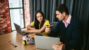 Ein Mentor und eine Schülerin sitzen an einem Tisch mit Laptops und einem kleinen Roboter und besprechen gemeinsam ein technisches Projekt.