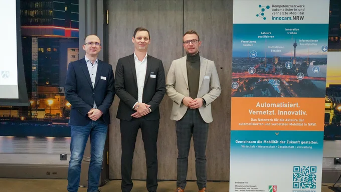 Drei Männer in Business-Kleidung stehen vor einer Tür neben einem Roll-up mit dem Titel 'innocam.NRW' und dem Slogan 'Automatisiert. Vernetzt. Innovativ.'