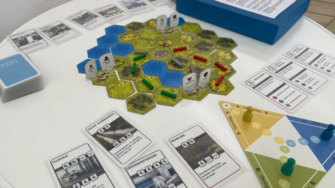 Auf einem weißen Stehstisch liegt das Wabenförmige Spielbrett zum Spiel "Landpartie" sowie das dreieckige Layerboard. Drumherum liegen spielkarten.