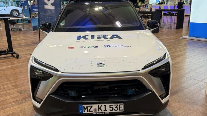 Weißes autonomes Fahrzeug mit KIRA-Logo auf einer Messeausstellung, frontal aufgenommen.