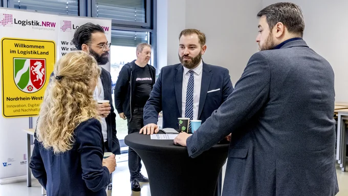 Mehrere Personen in Businesskleidung stehen um einen hohen Stehtisch in einem hellen Raum mit großen Fenstern. Im Hintergrund ein gelbes Schild mit dem Text 'Willkommen im LogistikLand Nordrhein-Westfalen'.