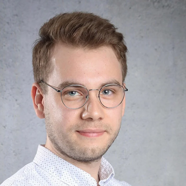 Porträt von Tim Rehbronn mit Brille, kurzen braunen Haaren in einem weißen Hemd vor grauem Hintergrund.