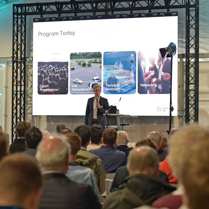 „Eine Person hält eine Präsentation vor einem Publikum in einem Konferenzzelt. Auf der großen Leinwand hinter ihr steht ‚Program Today‘ mit vier Themen: Expert Presentations, Live Demonstrations, Technical Exhibition und Networking. Die Veranstaltung hat ein professionelles Umfeld mit Beleuchtung und Bühnenaufbau.