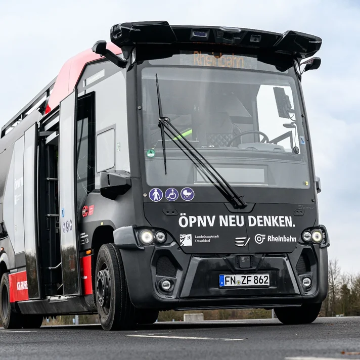 Vorderansicht des gleichen autonomen Shuttlebusses mit geöffneter Tür. Beschriftung „ÖPNV neu denken“ und Symbole für barrierefreie Zugänge sichtbar.
