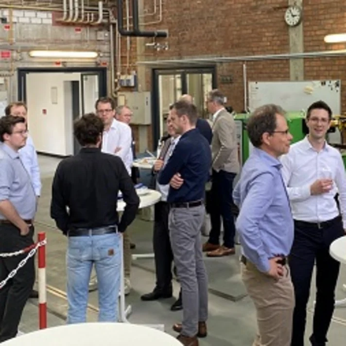 Fachleute tauschen sich in einer Werkhalle an Stehtischen aus; Maschinen und Technik im Hintergrund.