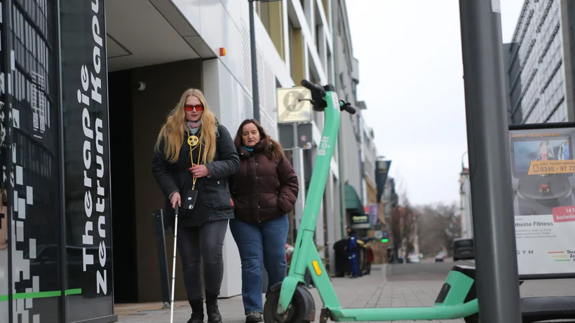 Blinde Frau mit Blindenstock und Begleitperson gehen auf einem Gehweg. Direkt vor ihnen steht ein abgestellter E-Scooter, der den Weg blockiert.