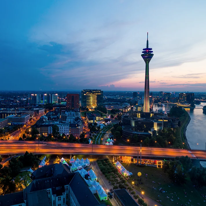 Ansicht der Stadt Düsseldorf aus der Vogelperspektive in der Dämmerung mit gelb-orange beleuchteten Straßen, blauem Farbton , dem Fluß Rhein auf der rechten Seite und einem Fernsehturm in der Mitte.