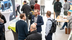 Mehrere Menschen unterhalten sich bei einer Konferenz im Foyer, während andere an Infoständen und Laptops arbeiten.