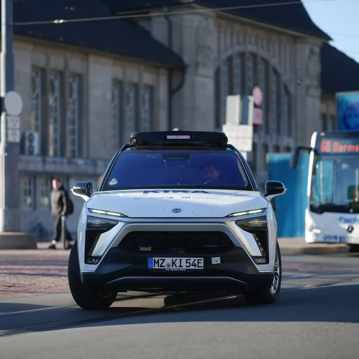 MAn sieht ein autonomes Fahrzeug von vorne, vone inem Bahnhof wegfahren, daneben ein Bus mit der Aufschrift "Darmstadt"