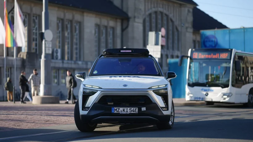 MAn sieht ein autonomes Fahrzeug von vorne, vone inem Bahnhof wegfahren, daneben ein Bus mit der Aufschrift "Darmstadt"