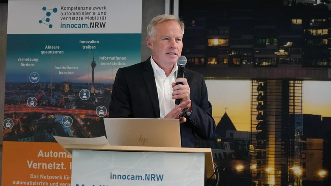 Mann in Anzug steht hinter einem Rednerpult mit Mikrofon, im Hintergrund Banner und Schild mit Aufschrift 'innocam.NRW Mobility Meeting 2025'.