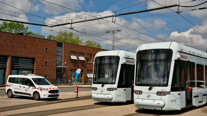 Zwei moderne Straßenbahnen und ein weißes Dienstfahrzeug mit roten Warnmarkierungen auf einem Betriebshof vor einem Backsteingebäude unter blauem Himmel mit Wolken