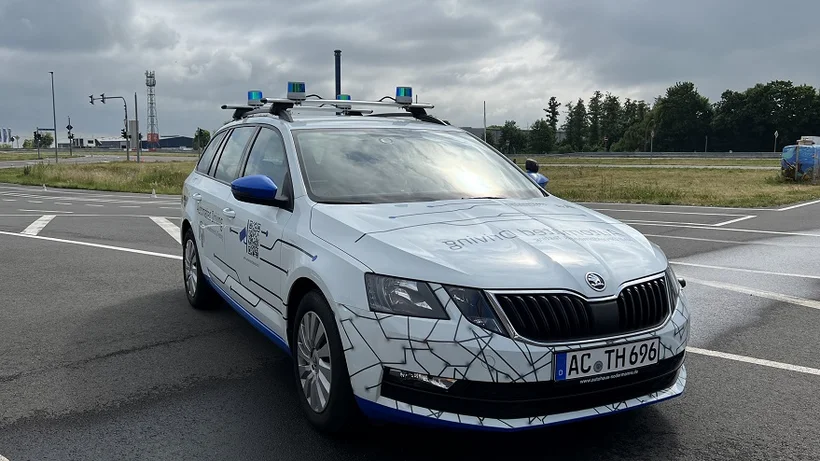 Autonomes Forschungsfahrzeug auf Straße mit Sensorik auf dem Dach. Es ist weiß mit schwarzer Linienmusterung.