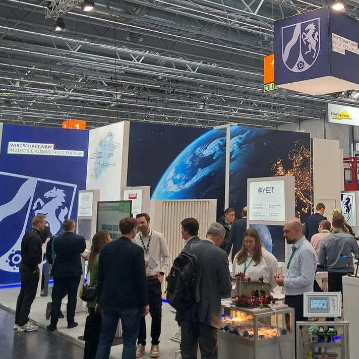 Messestand mit mehreren Personen, großen Bildschirmen und Informationsständen in einer Halle mit sichtbarer Decke und Beleuchtung