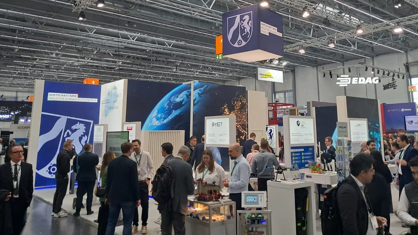 Messestand mit mehreren Personen, großen Bildschirmen und Informationsständen in einer Halle mit sichtbarer Decke und Beleuchtung