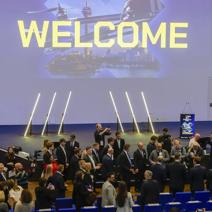 Große Gruppe von Menschen in einem Auditorium mit der Projektion 'WELCOME' und 'KEYNOTES' auf der Wand