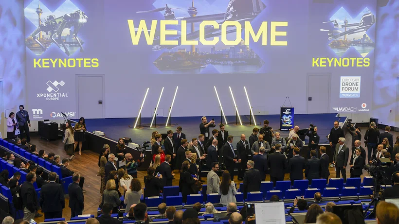 Große Gruppe von Menschen in einem Auditorium mit der Projektion 'WELCOME' und 'KEYNOTES' auf der Wand