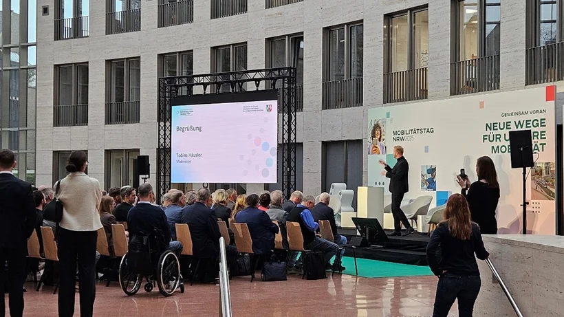 Im Atrium eines modernen Gebäudes spricht ein Mann auf einer Bühne vor einem Publikum, das teils sitzt, teils steht, bei der Veranstaltung Mobilitätstag NRW 2025 in Düsseldorf.