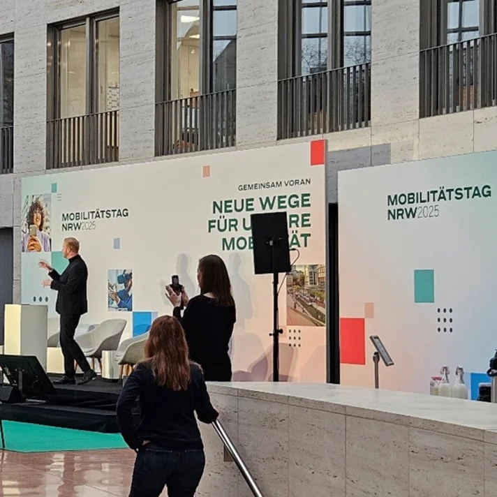 Im Atrium eines modernen Gebäudes spricht ein Mann auf einer Bühne vor einem Publikum, das teils sitzt, teils steht, bei der Veranstaltung Mobilitätstag NRW 2025 in Düsseldorf.