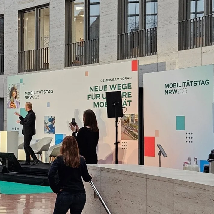 Im Atrium eines modernen Gebäudes spricht ein Mann auf einer Bühne vor einem Publikum, das teils sitzt, teils steht, bei der Veranstaltung Mobilitätstag NRW 2025 in Düsseldorf.