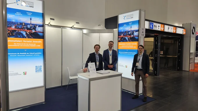 Drei Männer stehen am Messestand von innocam.NRW auf der XPONENTIAL 2025 und präsentieren Informationsmaterial zur vernetzten Mobilität.