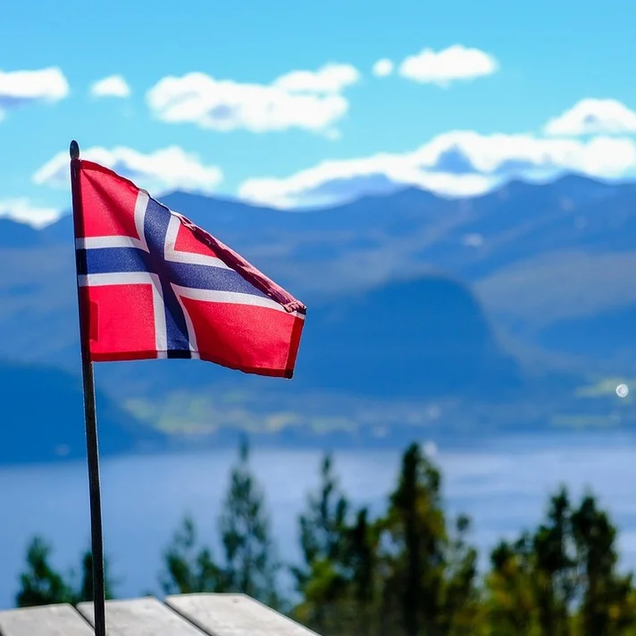 Norwegische Flagge auf einem Fahnenmast vor Berg- und Seelandschaft mit blauem Himmel und Wolken.