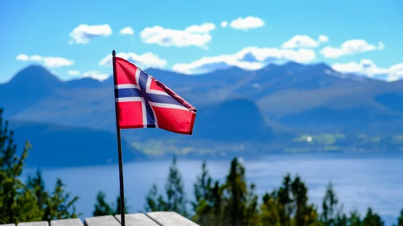 Norwegische Flagge auf einem Fahnenmast vor Berg- und Seelandschaft mit blauem Himmel und Wolken.