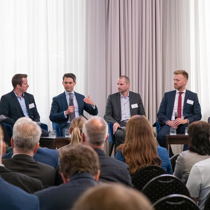Fünf Personen sitzen auf einer Bühne in einem Podiumsgespräch vor Publikum, zwei Personen haben verschwommene Gesichter, einer spricht und gestikuliert mit der Hand.