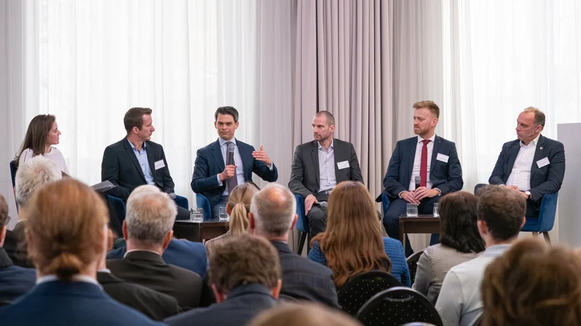 Fünf Personen sitzen auf einer Bühne in einem Podiumsgespräch vor Publikum, zwei Personen haben verschwommene Gesichter, einer spricht und gestikuliert mit der Hand.