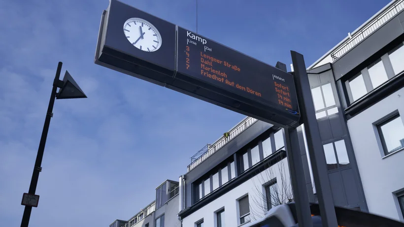 Elektronisches Fahrgastinformationsdisplay mit Abfahrtszeiten und integrierter Uhr an einer Bushaltestelle in der Stadt, vor einem Wohngebäude und blauem Himmel.