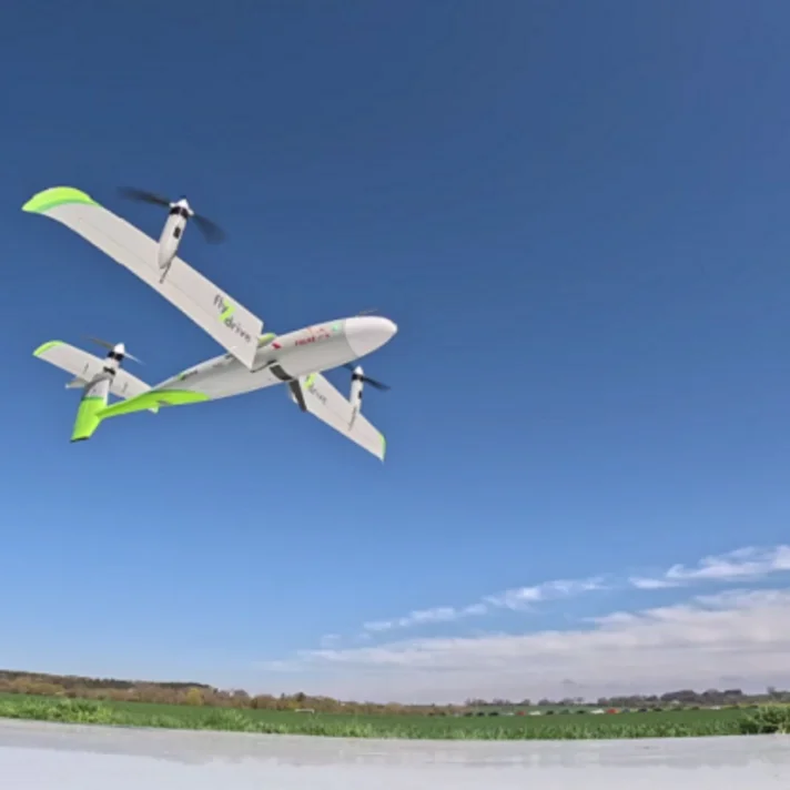 Weiß-grüne senkrechtstartende Drohne (VTOL) im Horizontalflug über einem landwirtschaftlichen Feld, klarer blauer Himmel im Hintergrund.