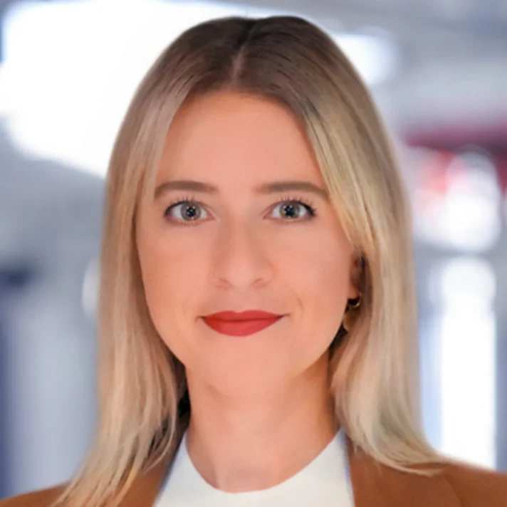 Porträt von Cindy Pöschk, Referentin für Öffentlichkeitsarbeit, mit blondem Haar, rotem Lippenstift und braunem Blazer, vor verschwommenem Hintergrund.