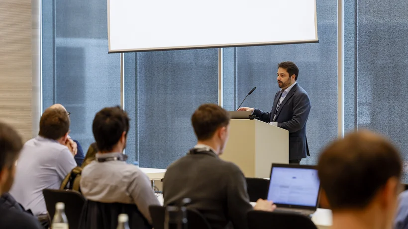 Mann im Anzug steht an einem Rednerpult vor einem Projektionsschirm und spricht zu mehreren sitzenden Zuhörern in einem Konferenzraum