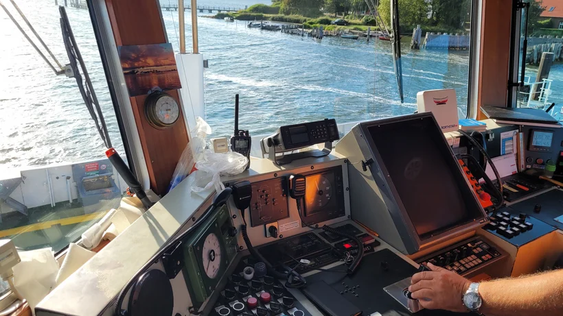 Innenansicht des Steuerstands einer Fähre mit verschiedenen nautischen Instrumenten und Displays, eine Hand am Steuerhebel, Wasser und Uferlandschaft sichtbar durch die Fenster.