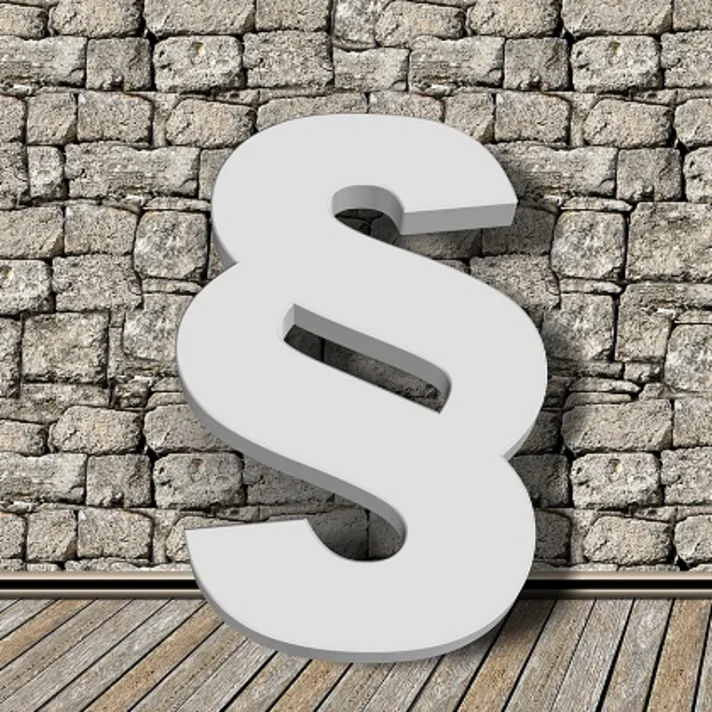 Illustration eines großen, dreidimensionalen Paragrafenzeichens auf einem Parkettboden, gegen eine Steinwand gelehnt und darauf einen Schatten werfend..