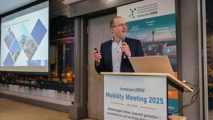 Mann steht am Rednerpult mit Mikrofon und Fernbedienung, im Hintergrund Präsentationsfolie und Banner mit inncam.NRW-Logo und Text zum Mobility Meeting 2025.