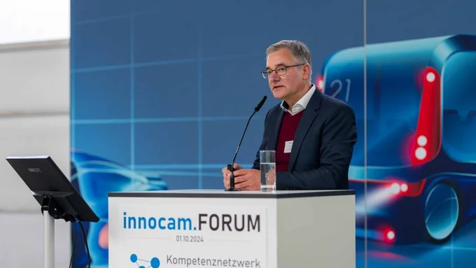 Ein Mann spricht am Rednerpult mit „innocam.FORUM“-Logo vor einer blauen Wand mit futuristischem Fahrzeugmotiv.