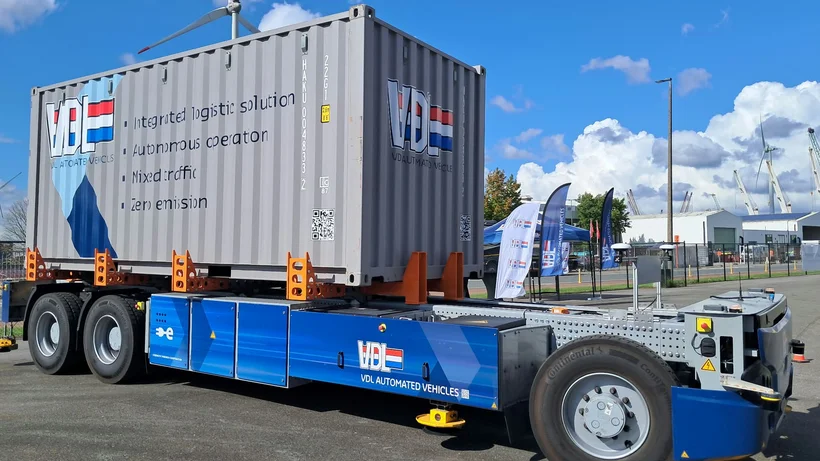 Autonom fahrendes, blaues Fahrzeug mit aufgeladenem grauem Container und Aufschrift 'VDL AUTOMATED VEHICLES' auf einem Industrieparkplatz