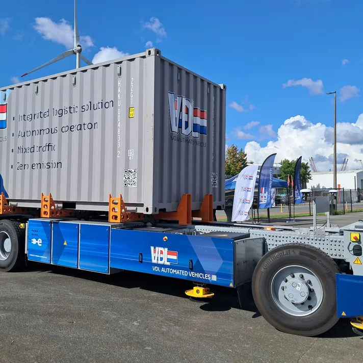 Autonom fahrendes, blaues Fahrzeug mit aufgeladenem grauem Container und Aufschrift 'VDL AUTOMATED VEHICLES' auf einem Industrieparkplatz