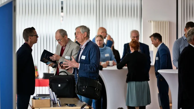 Mehrere Personen in Businesskleidung unterhalten sich in einem hellen Raum mit Stehtischen und Vorhängen im Hintergrund