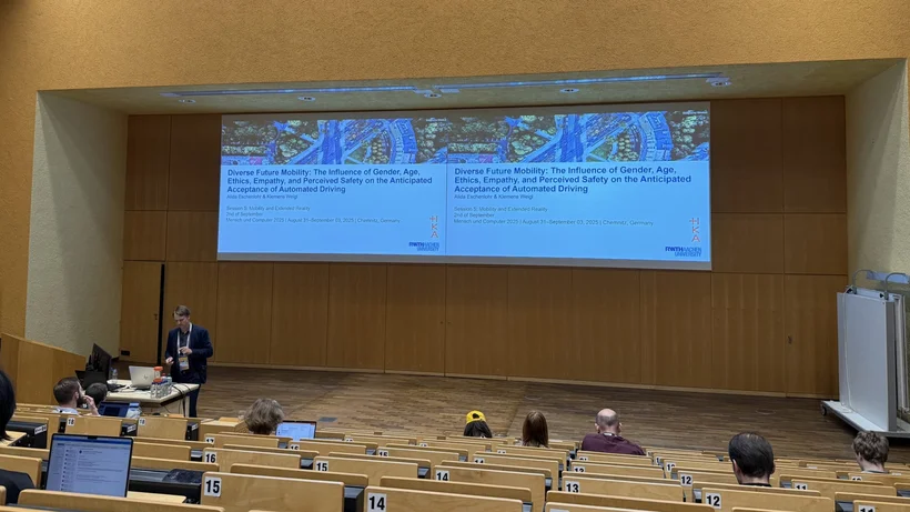 Auf der Bühne eines Hörsaals mit Zuschauern steht ein Mann hinter einem Tisch mit Laptop und präsentiert einen Vortrag, die Projektion hinter ihm zeigt eine Folienpräsentation.