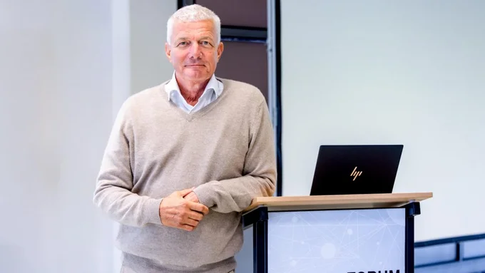 Mann mit hellgrauem Pullover und weißem Hemd lehnt an einem Rednerpult mit Laptop und Schriftzug 'innocam.FORUM 18.09.2025'.