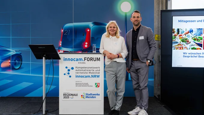 Zwei Personen posieren lächelnd am Rednerpult des innocam.FORUM mit Info-Bildschirm im Hintergrund.