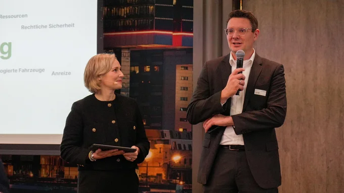 Zwei Personen stehen vor einer Präsentationswand, eine hält ein Mikrofon, die andere ein Tablet, beide tragen formelle Kleidung.