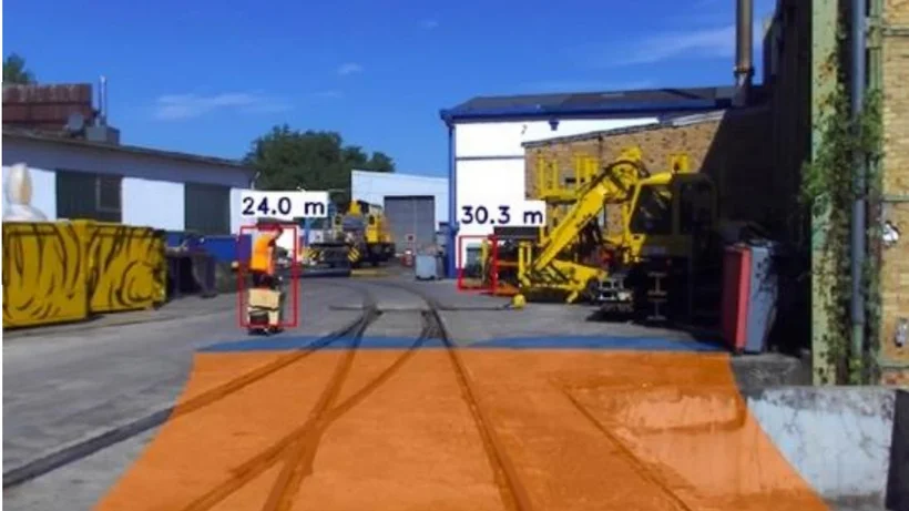 Industriegelände mit Gleisen, zwei gelb-orangefarbenen Baumaschinen und Entfernungsangaben in Metern in roten Rechtecken.