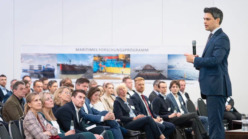 Mann im Anzug hält Mikrofon und spricht vor sitzendem Publikum in einem Konferenzraum mit Banner 'MARITIMES FORSCHUNGSPROGRAMM' im Hintergrund