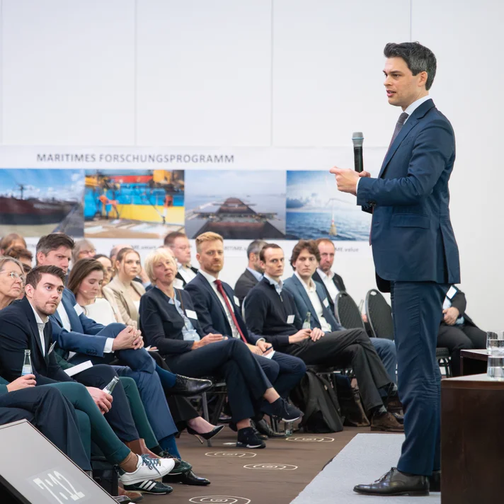 Mann im Anzug hält Mikrofon und spricht vor sitzendem Publikum in einem Konferenzraum mit Banner 'MARITIMES FORSCHUNGSPROGRAMM' im Hintergrund