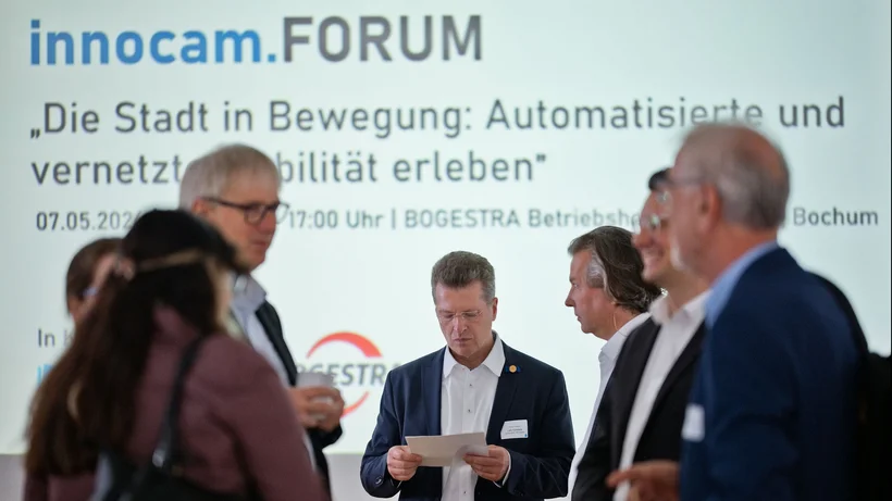 Gruppe von Personen in Businesskleidung vor einer Präsentationswand mit dem Schriftzug 'innocam.FORUM' und dem Thema 'Die Stadt in Bewegung Automatisierte und vernetzte Mobilität erleben'