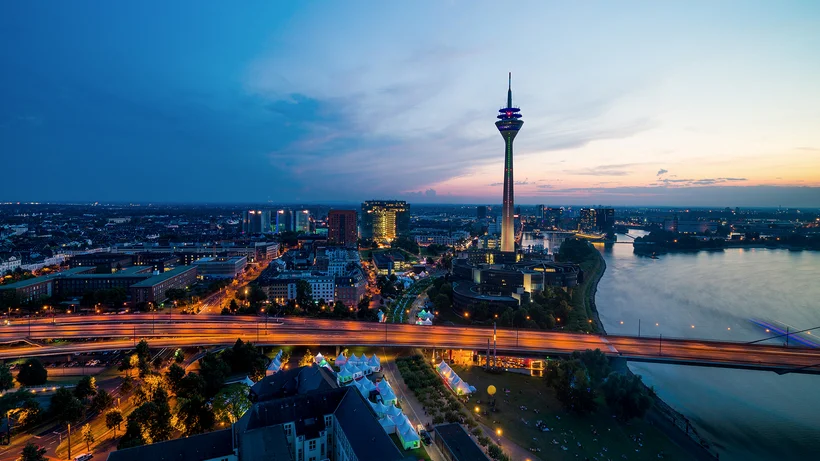 Ansicht der Stadt Düsseldorf aus der Vogelperspektive in der Dämmerung mit gelb-orange beleuchteten Straßen, blauem Farbton , dem Fluß Rhein auf der rechten Seite und einem Fernsehturm in der Mitte.
